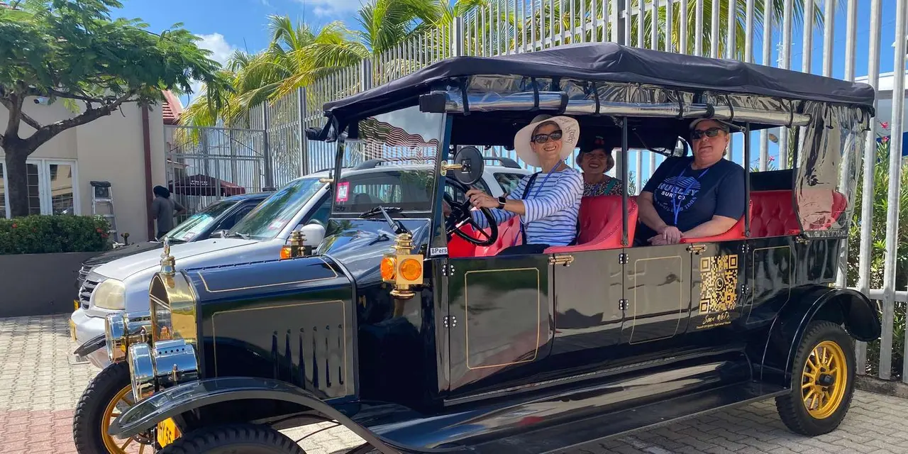 Two passengers in an open black Model T replica parked outside a white fence