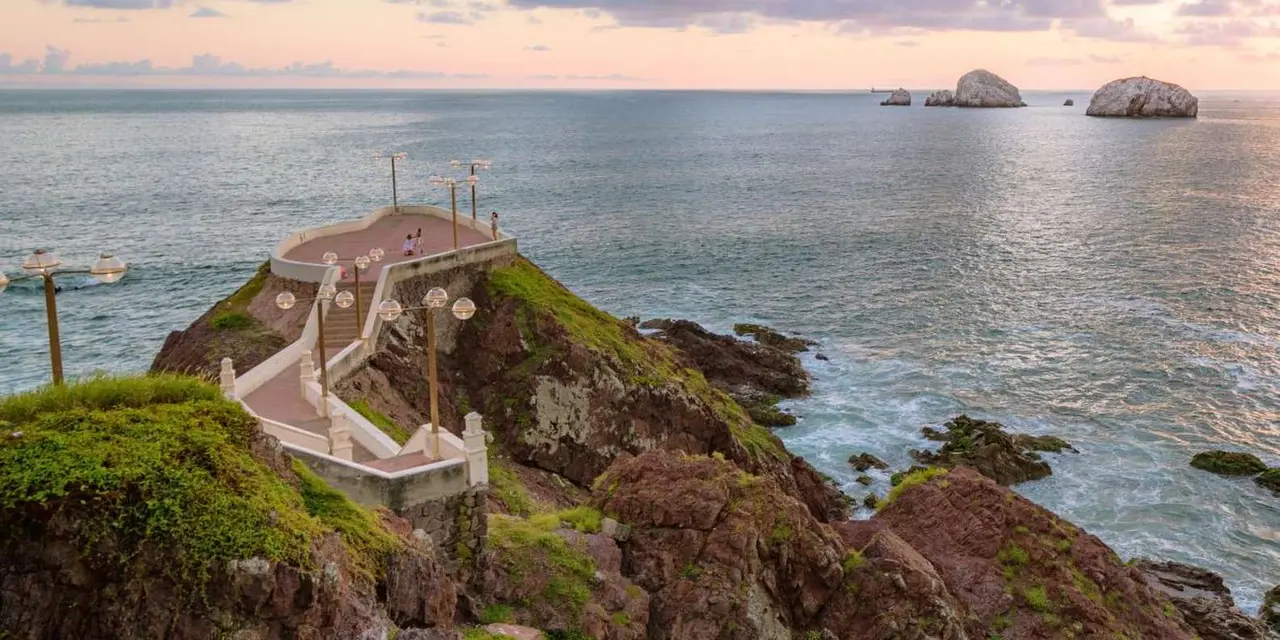 A curved stone viewing platform with lamp posts on a rocky green headland above the sea, with rock islands on the horizon