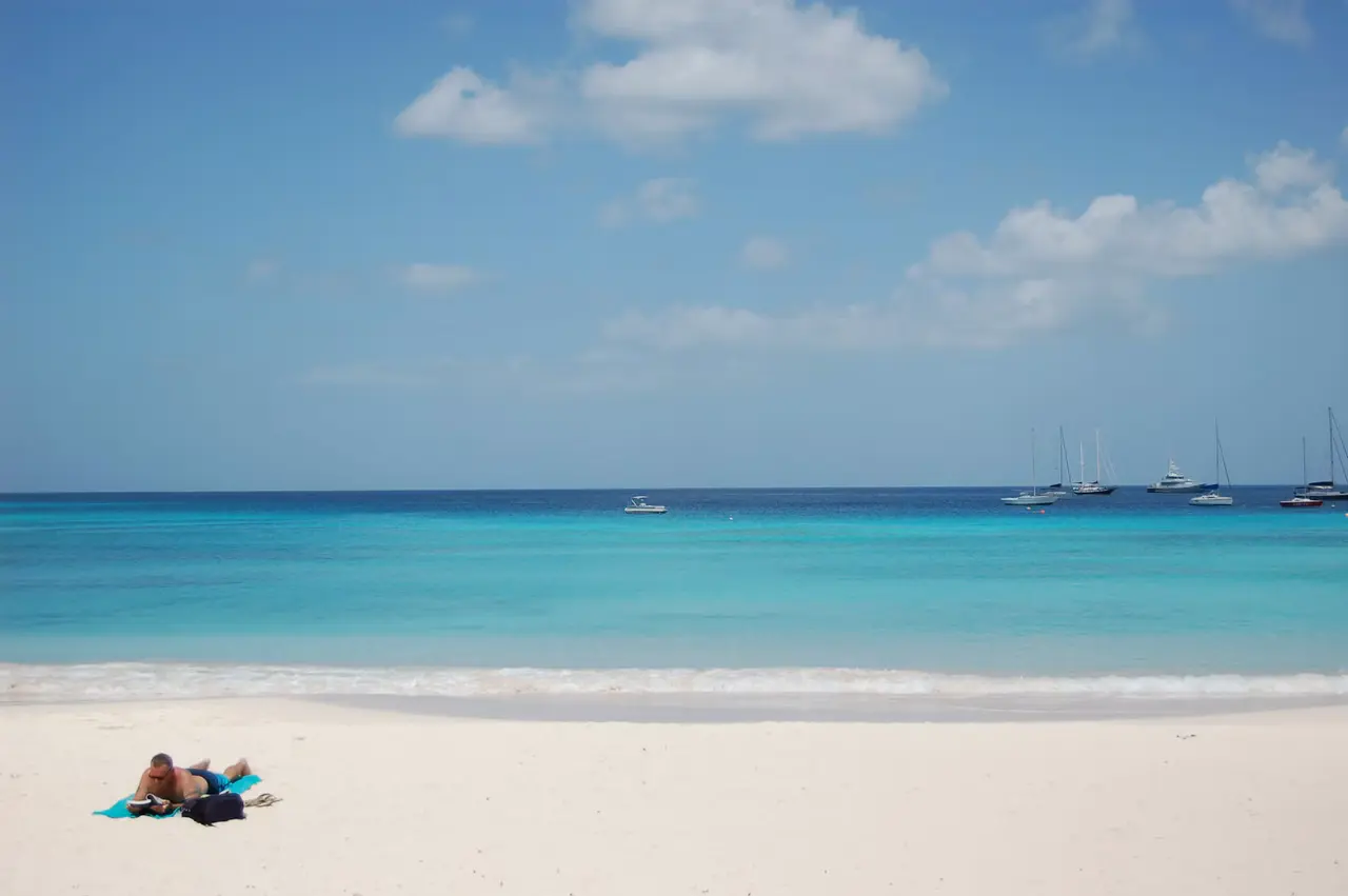 Turquoise Barbados water and a white sand beach under a blue sky with scattered clouds, a sunbather on a towel in the foreground and sailboats anchored offshore