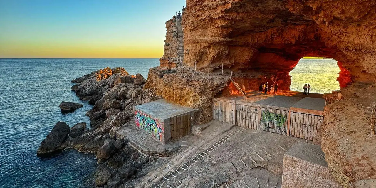 A sea cave with weathered boathouse doors on a rocky shore at sunset