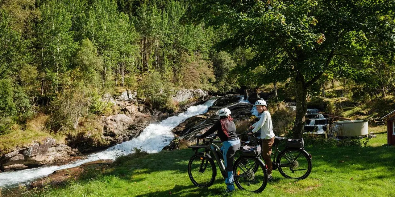 Two cyclists with e-bikes beside a stream and a small cascade