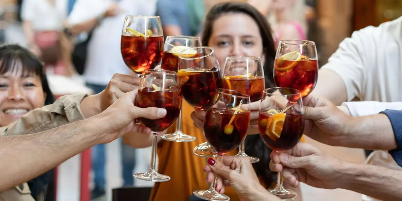 Hands clinking glasses of a red drink with orange slices, with smiling faces behind them