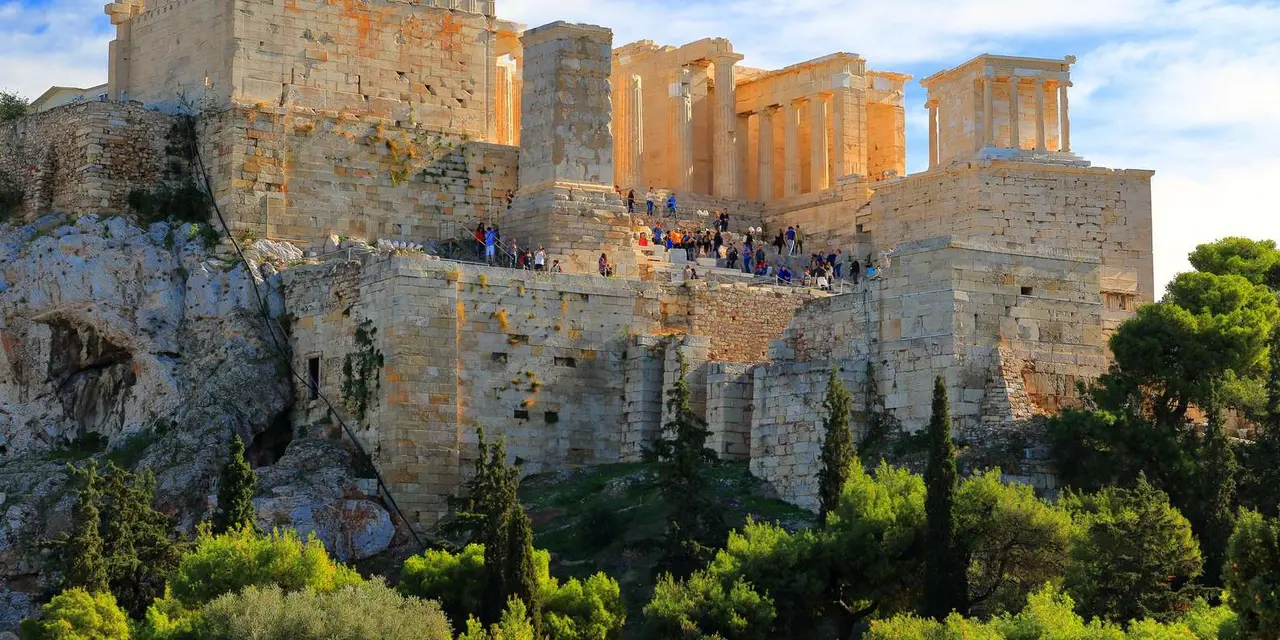 The Acropolis walls and its columned entrance rising above pine trees, visitors lined along the top