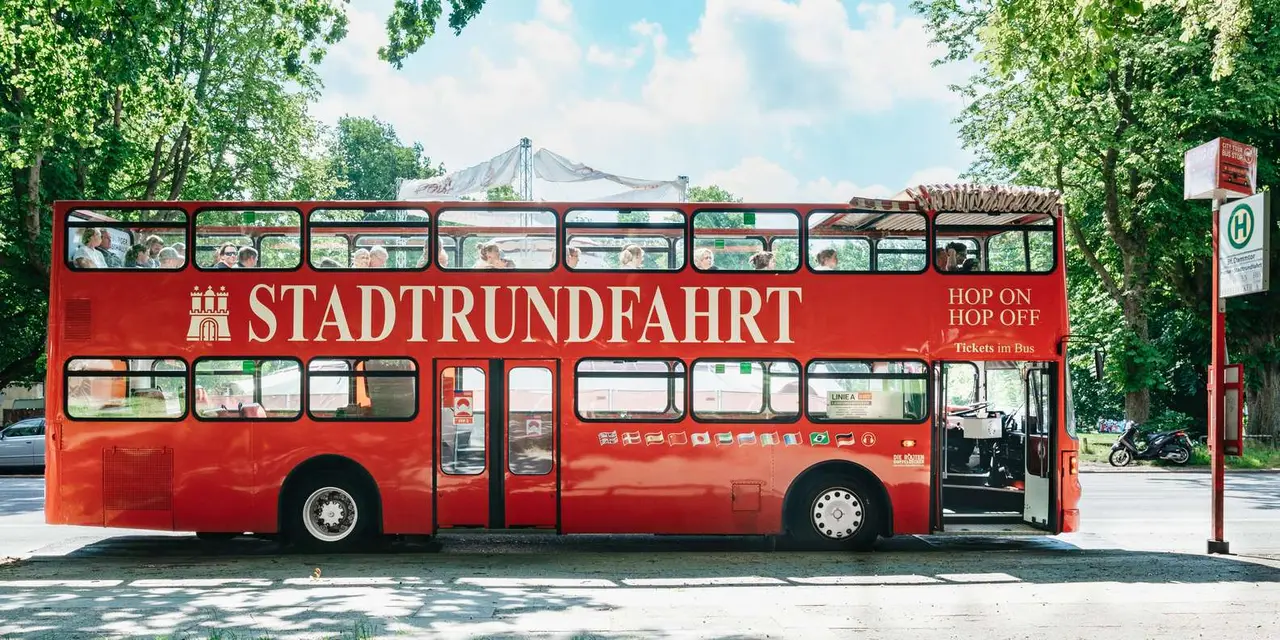 A red open-top double-decker bus marked Stadtrundfahrt Hop On Hop Off waiting at a tree-lined stop in Hamburg