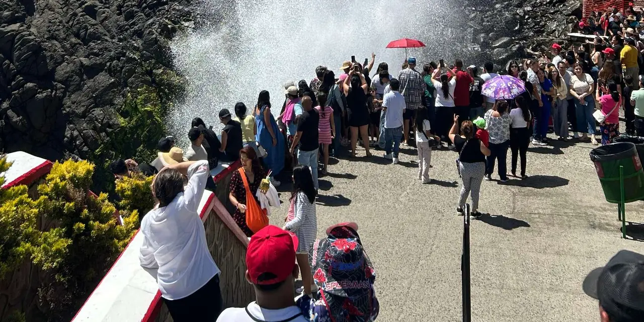 A crowd of visitors on a viewing walkway watching a tall plume of spray from the blowhole between dark rocks