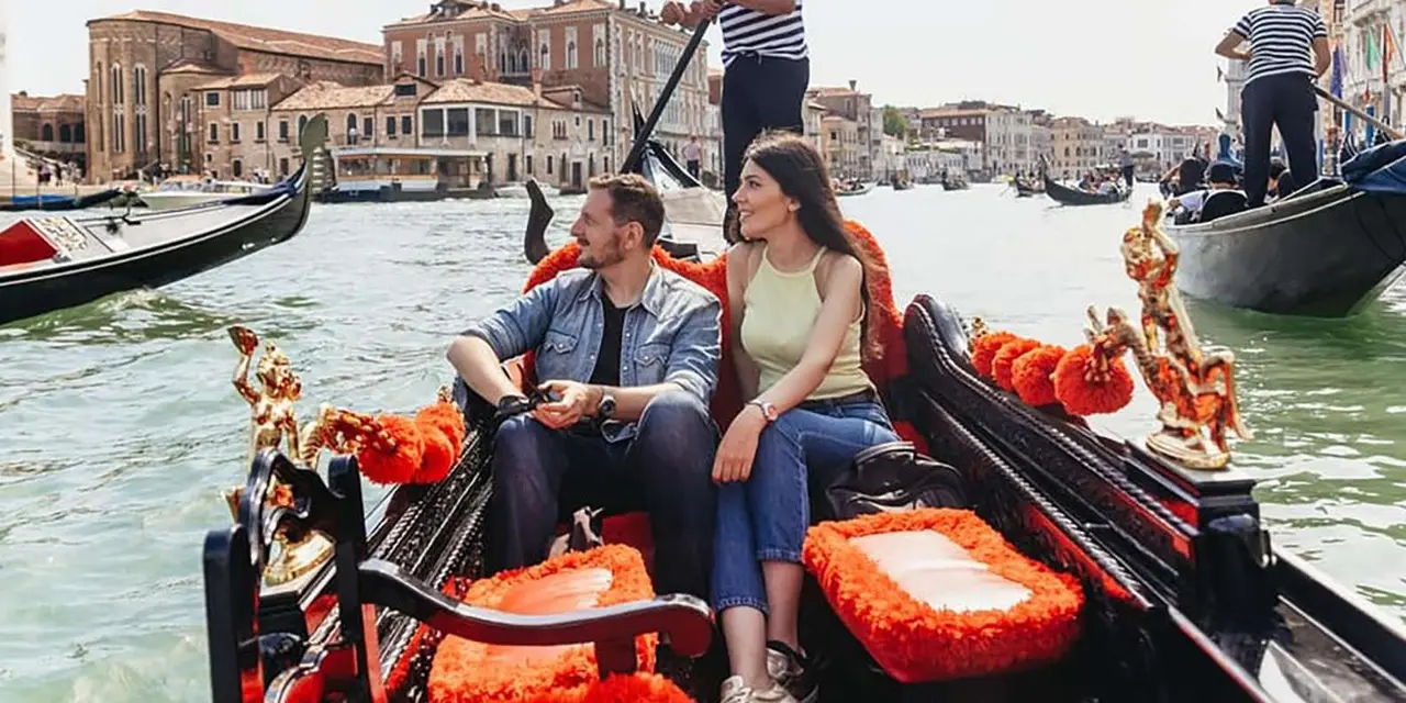 A couple sitting in a gondola on the Grand Canal with a gondolier standing at the stern and palazzi along the water behind them