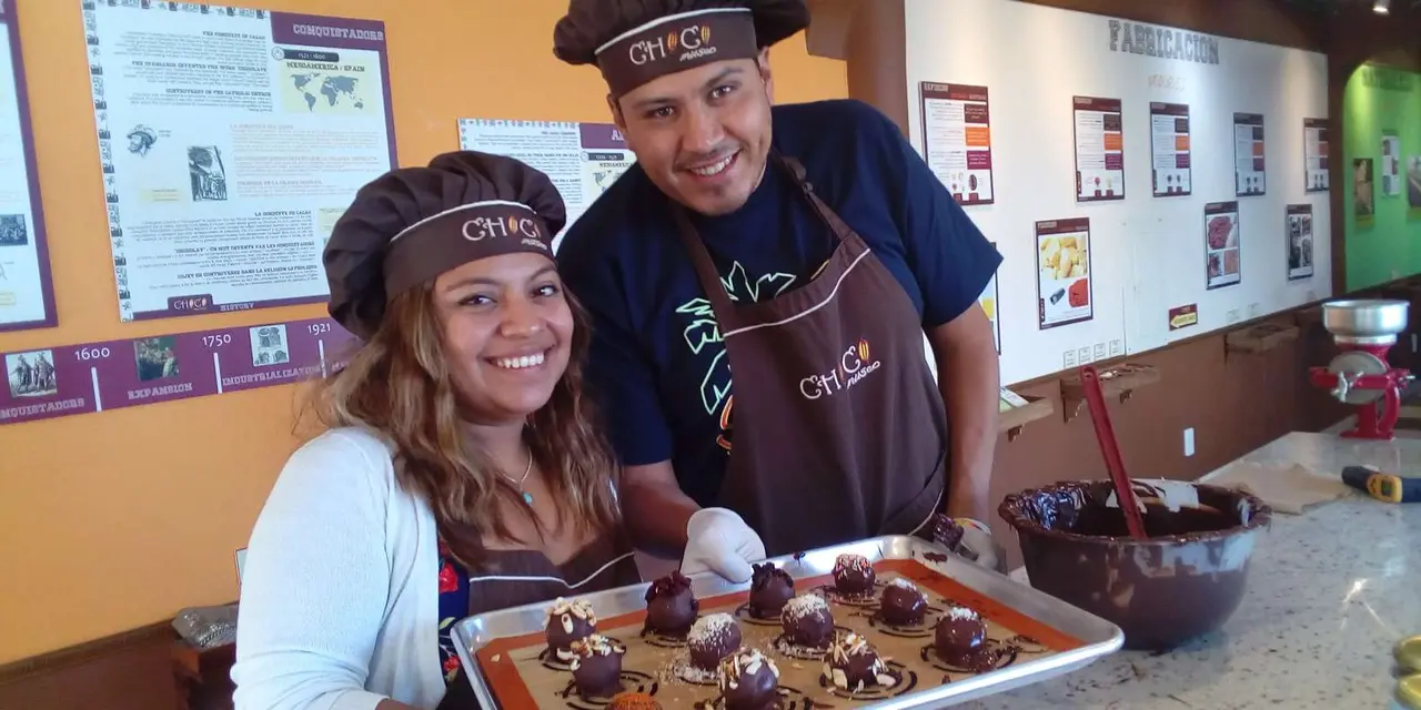 A smiling woman and man in aprons and caps holding a tray of freshly made chocolate truffles