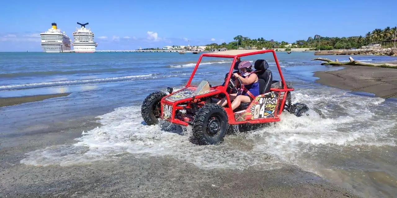 A red buggy with two helmeted riders crossing shallow water, with a cruise ship berthed behind it