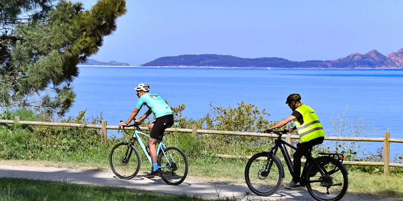 A cyclist in a helmet riding an electric bike along a coastal path above blue water, with hills across the bay