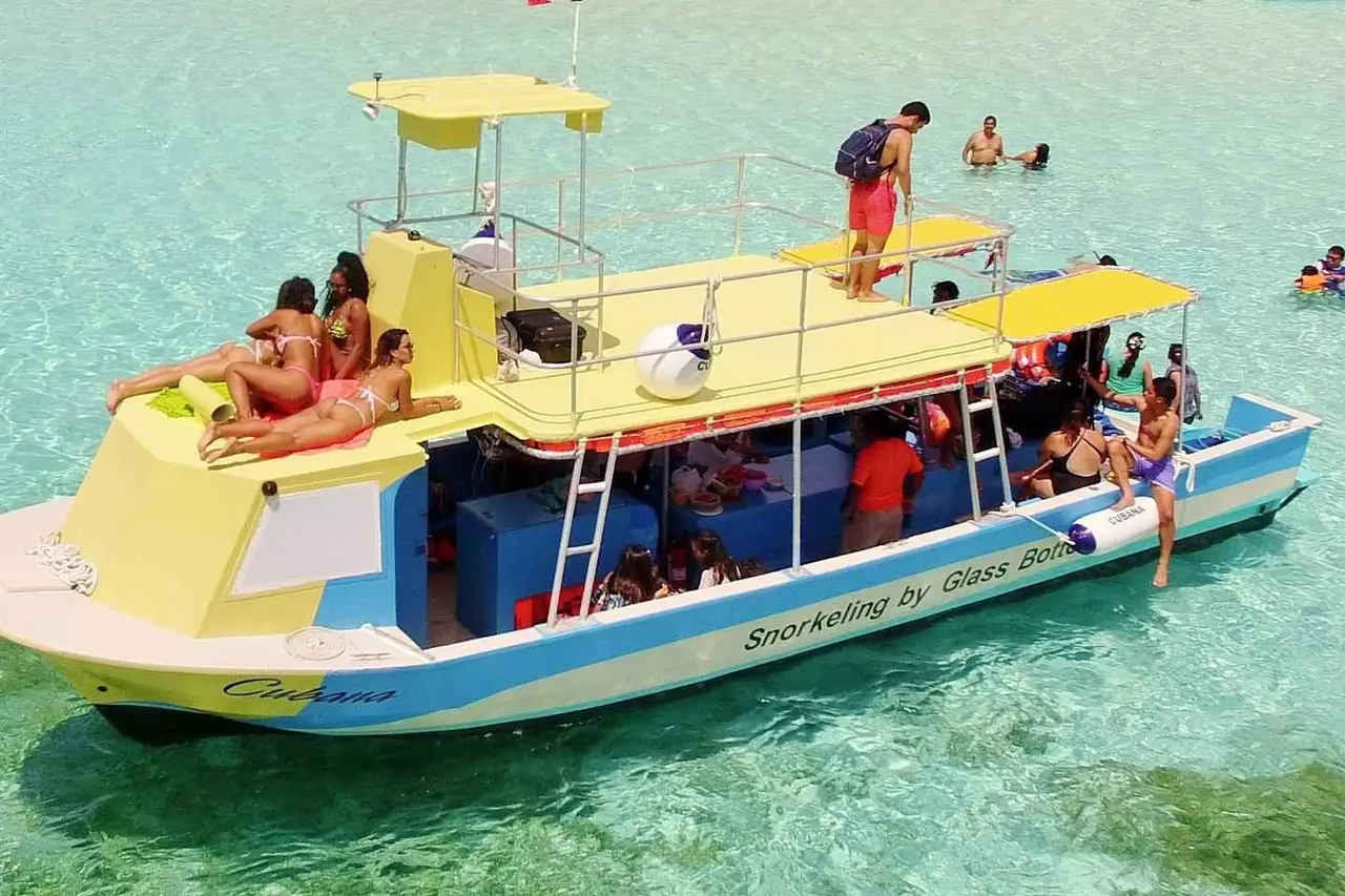 A yellow-and-blue two-deck glass-bottom boat anchored in shallow turquoise water, with passengers on the roof and swimmers nearby