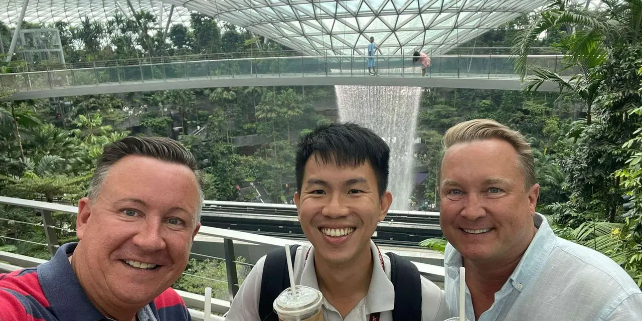 A guide and two visitors smiling for a photo under the glass dome and indoor waterfall at Jewel Changi Airport
