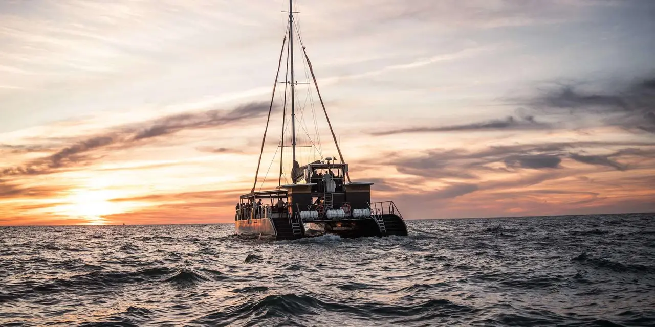 A two-masted catamaran carrying passengers on open water beneath a golden-orange sunset sky streaked with cloud