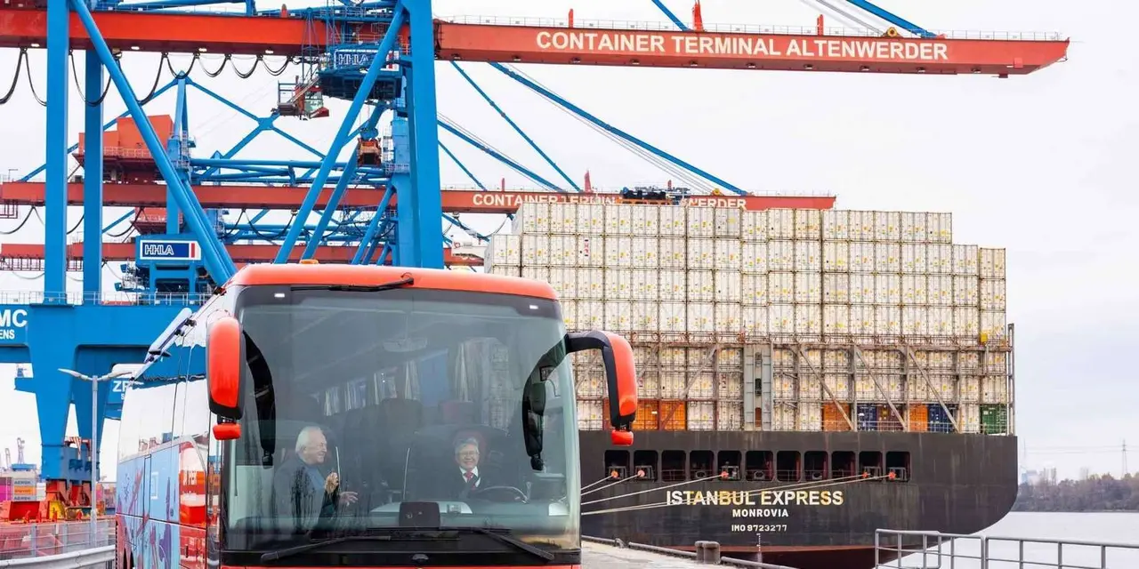 A red tour bus in front of tall blue container cranes and a stacked container ship at the Altenwerder container terminal