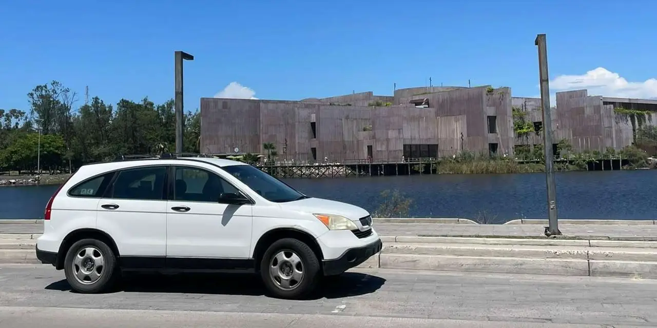 A white SUV parked on a roadside in front of a lagoon and a low brown building