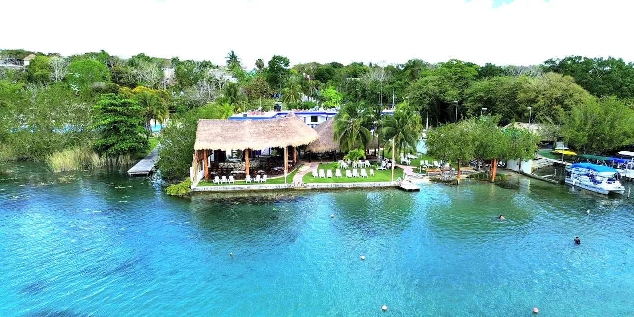 Aerial view of a thatched lakeside restaurant with a lawn of white chairs beside the bright turquoise water of Bacalar Lagoon, with pontoon boats moored at the right