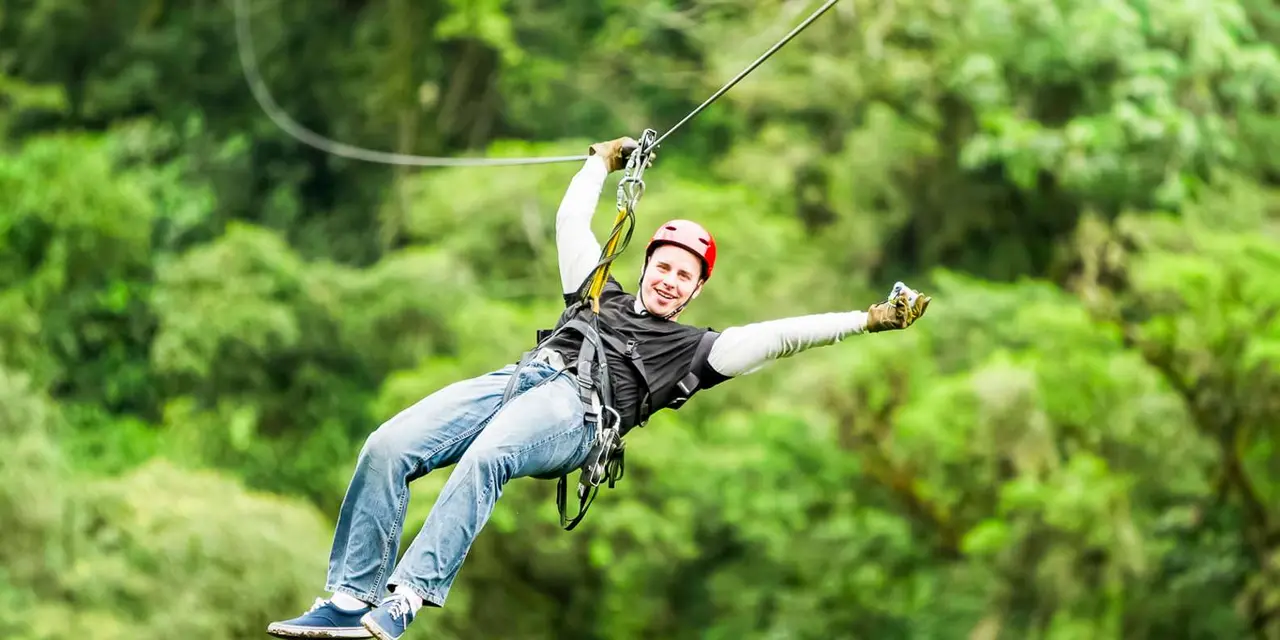 A man in a red helmet riding a zipline above green forest