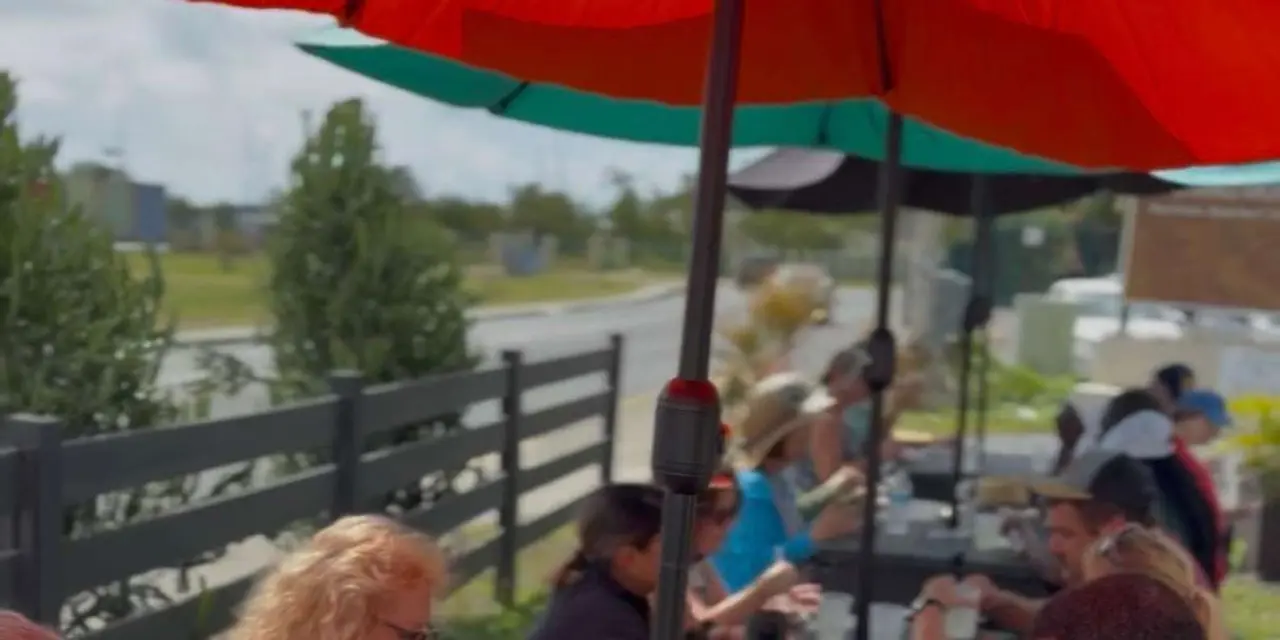 A group of visitors tasting food at an outdoor table under red and green umbrellas