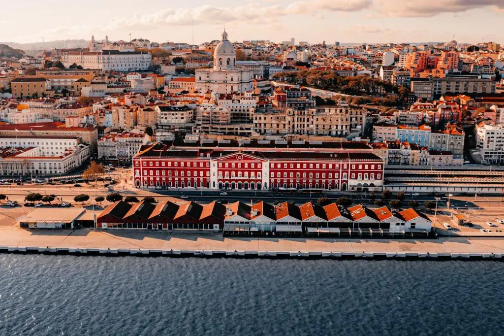 Aerial view of Santa Apolónia station's red-and-white facade on the Lisbon waterfront, where The Editory Riverside hotel occupies the building, with Alfama and the National Pantheon dome behind