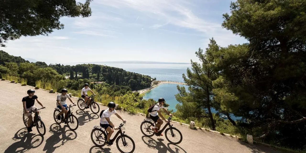 A line of cyclists riding a gravel path through trees with the sea below
