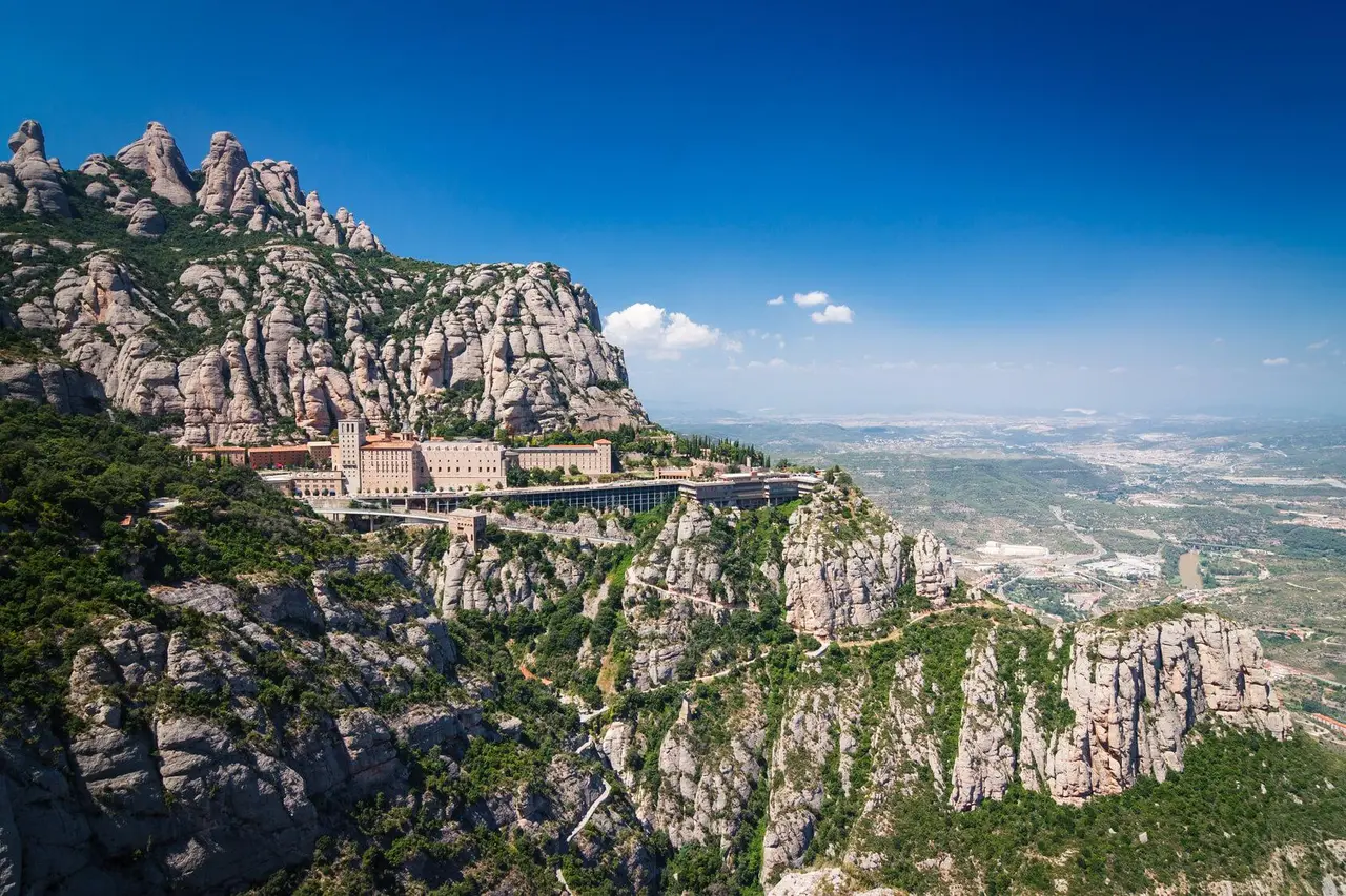 The monastery of Montserrat perched on a ledge below the jagged grey peaks, with green hills and a wide valley falling away under a deep blue sky