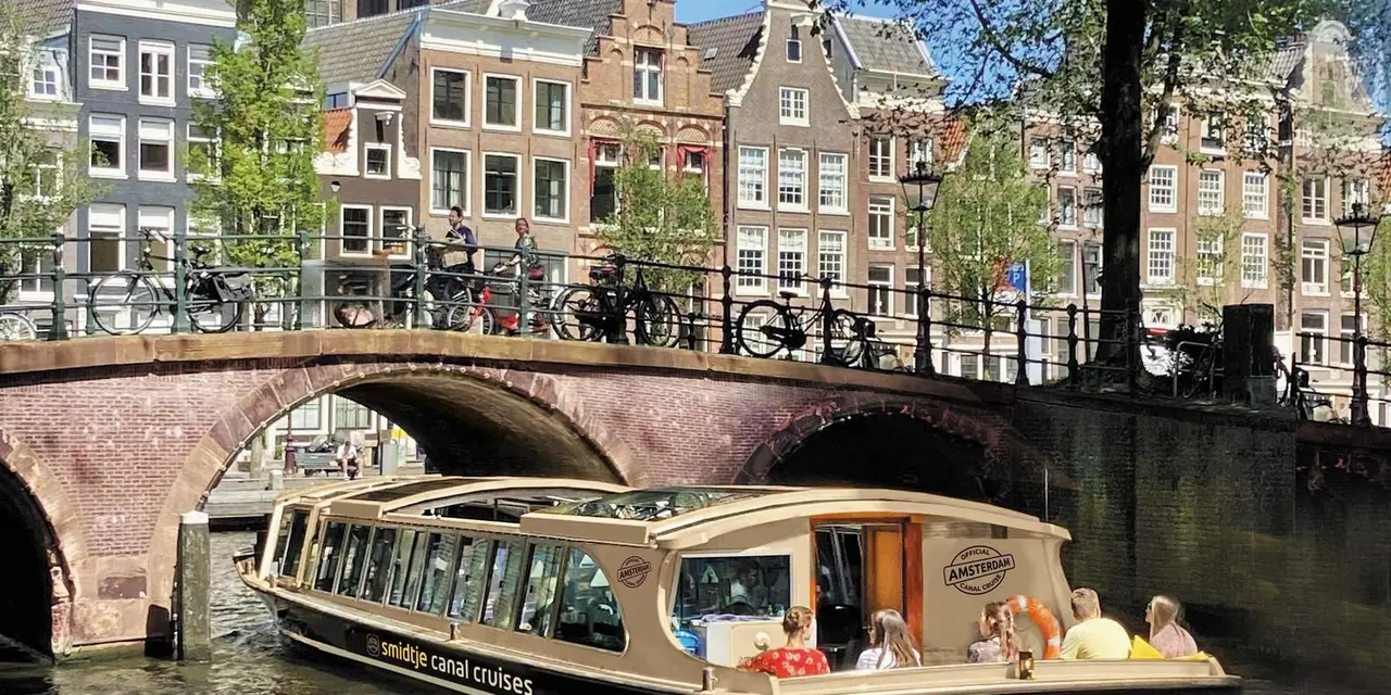 A cream and black glass-topped canal boat with passengers at the stern passing under a brick arch bridge lined with bicycles and tall canal houses