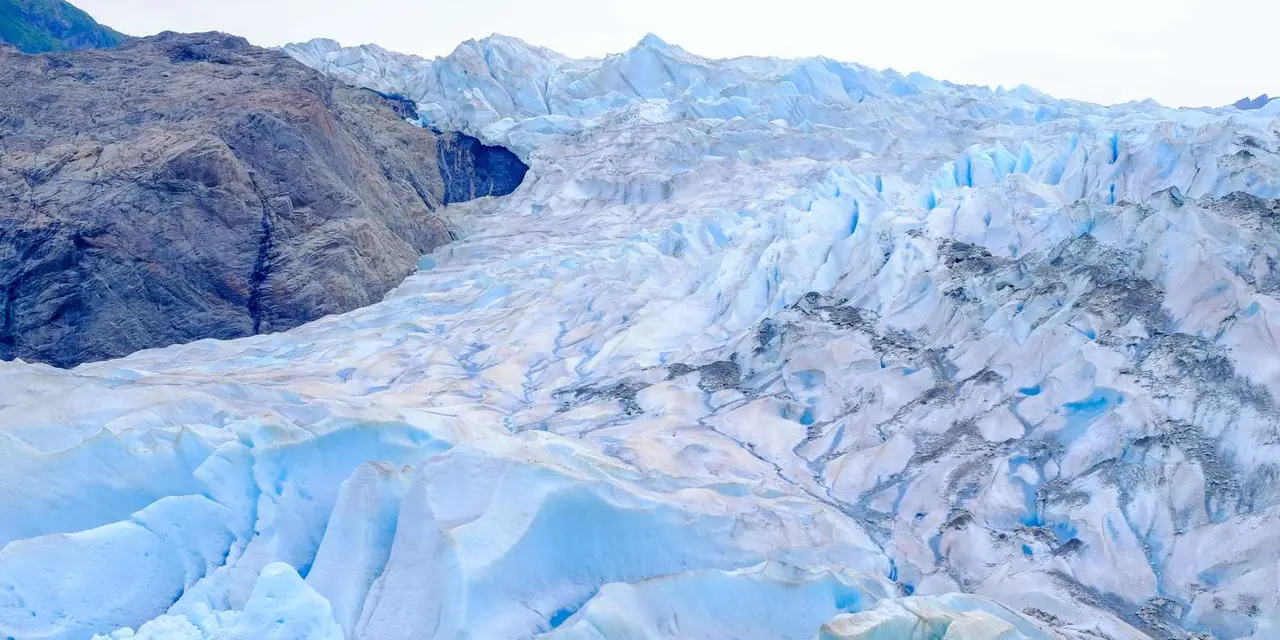 Close view of blue-white glacier ice with deep ridges and crevasses beside a dark rock wall
