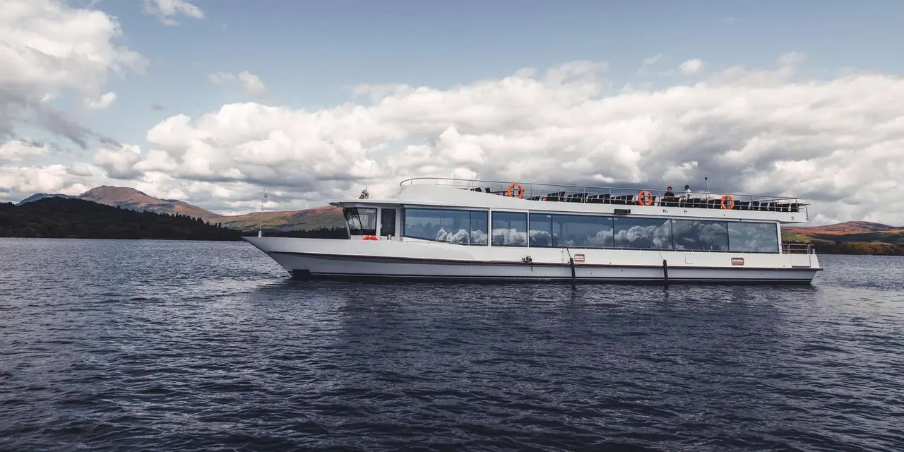 A white cruise boat with wide glass windows on dark loch water, low hills and a big cloudy sky behind