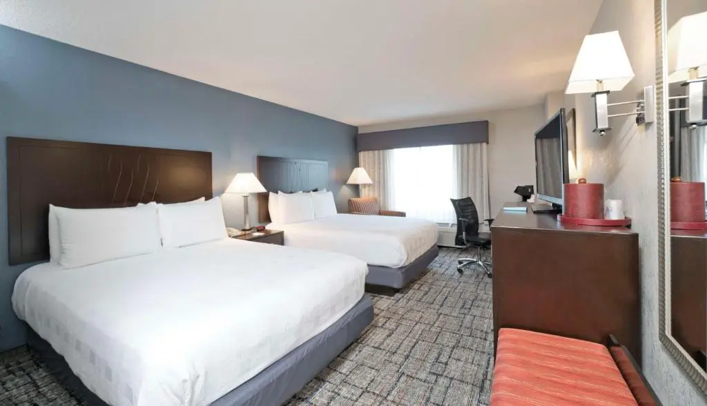 A Best Western Plus guest room with two queen beds, a slate-blue accent wall, a desk with a chair and a television