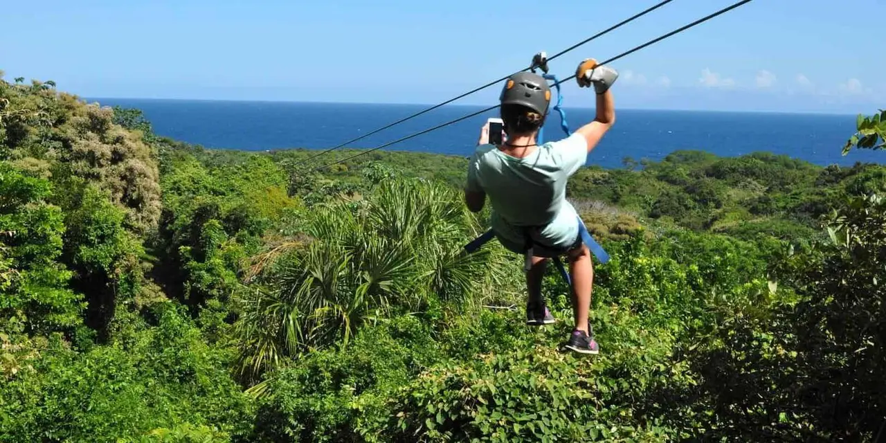 A zip-liner in a gray shirt and helmet crosses above green jungle with the blue sea on the horizon behind