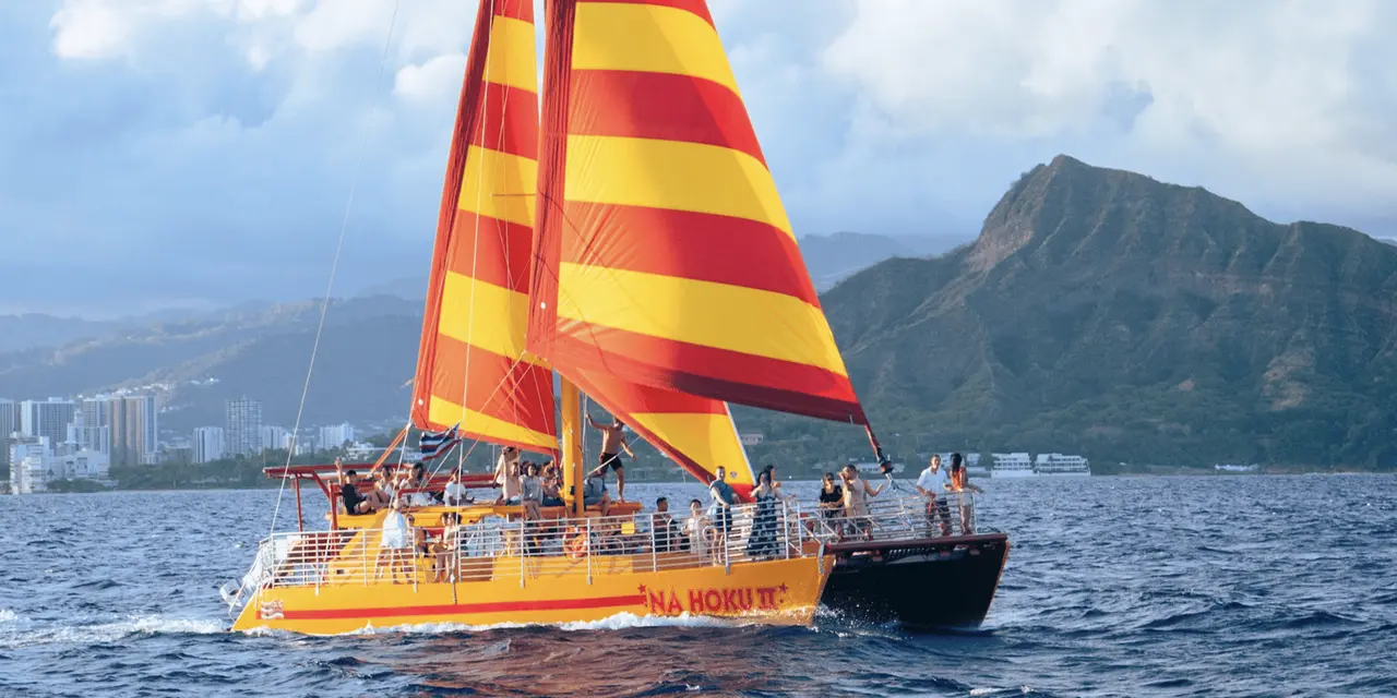 A yellow-and-red striped catamaran sail in the foreground with Diamond Head rising behind blue water