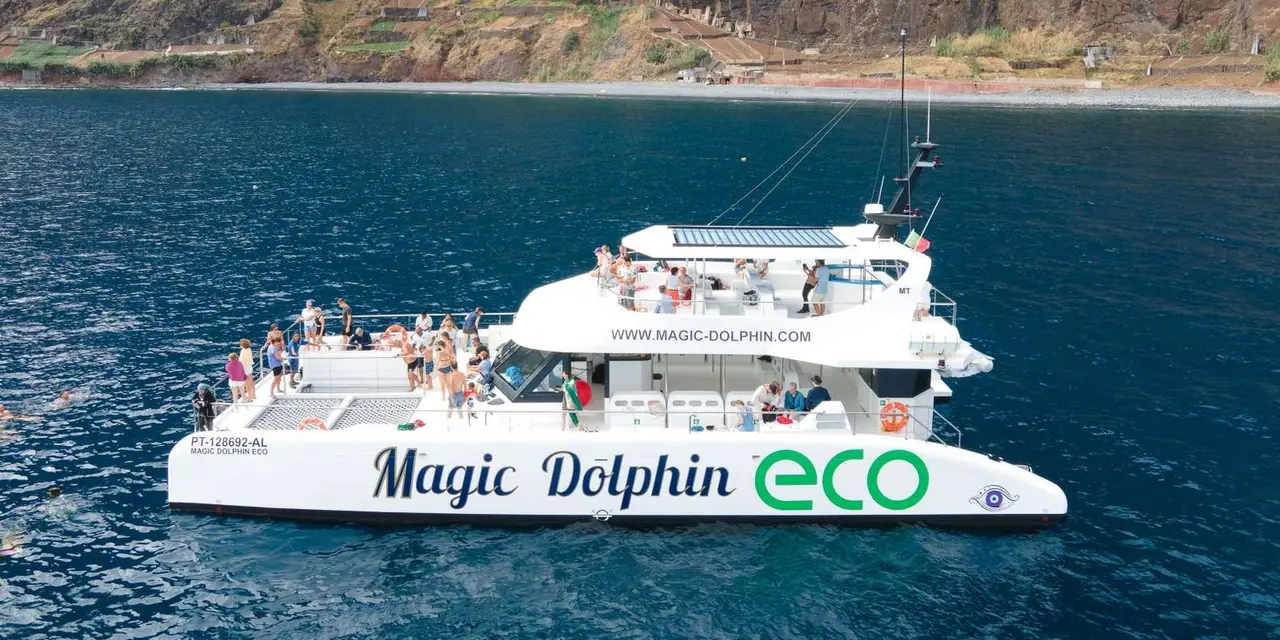 A white catamaran marked Magic Dolphin Eco carrying passengers on its decks, with a rocky cliff behind it on dark blue water