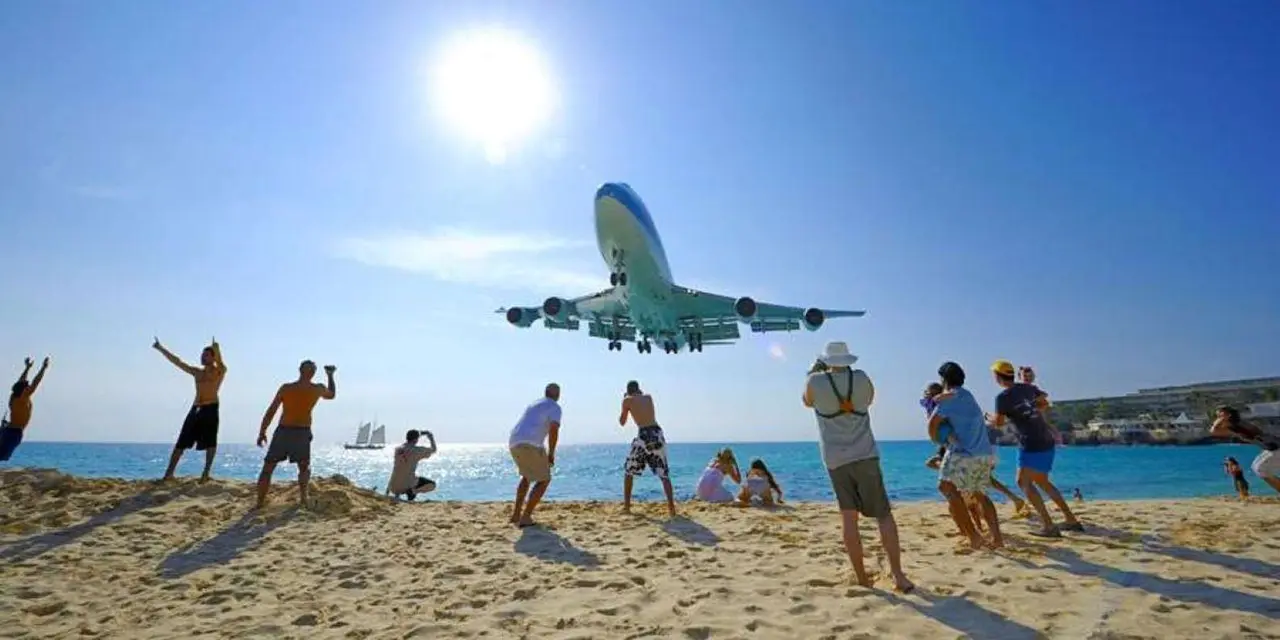 A jet landing low over a sunny Maho Beach, with a few people on the sand reaching up toward it