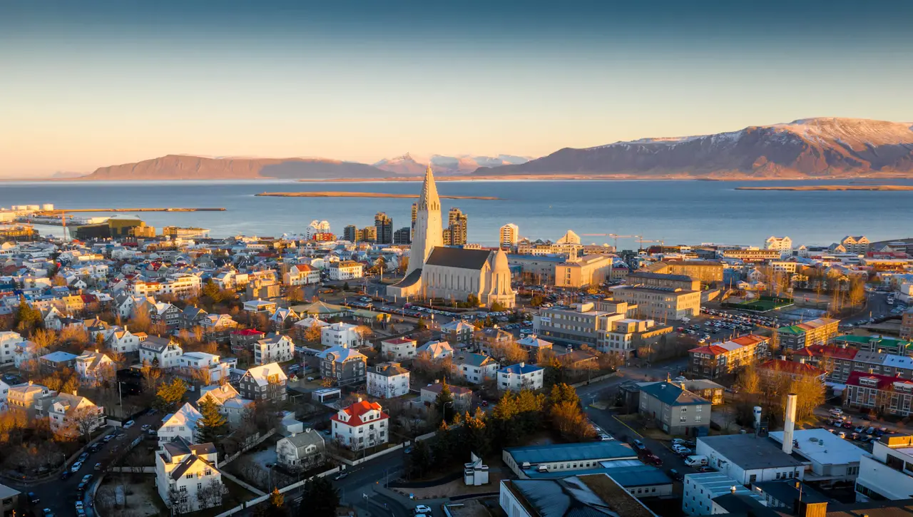 Aerial view of Reykjavik at sunrise, the Hallgrímskirkja tower rising above rows of rooftops with the bay and snow-dusted mountains glowing orange beyond