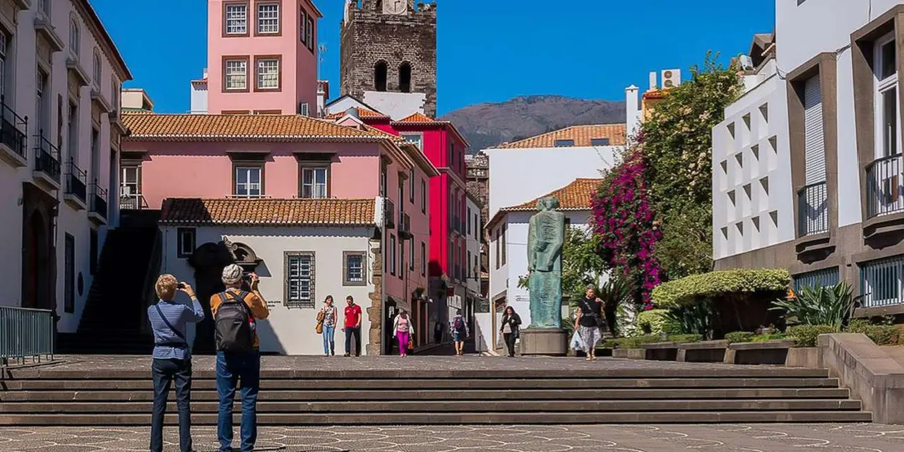 Pastel-coloured buildings and a cathedral bell tower around a sunny Funchal square with a statue, people stopping to take photos