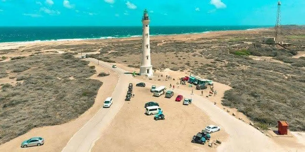 Aerial view of the white California Lighthouse tower on a sandy headland with turquoise sea behind and a few parked vehicles