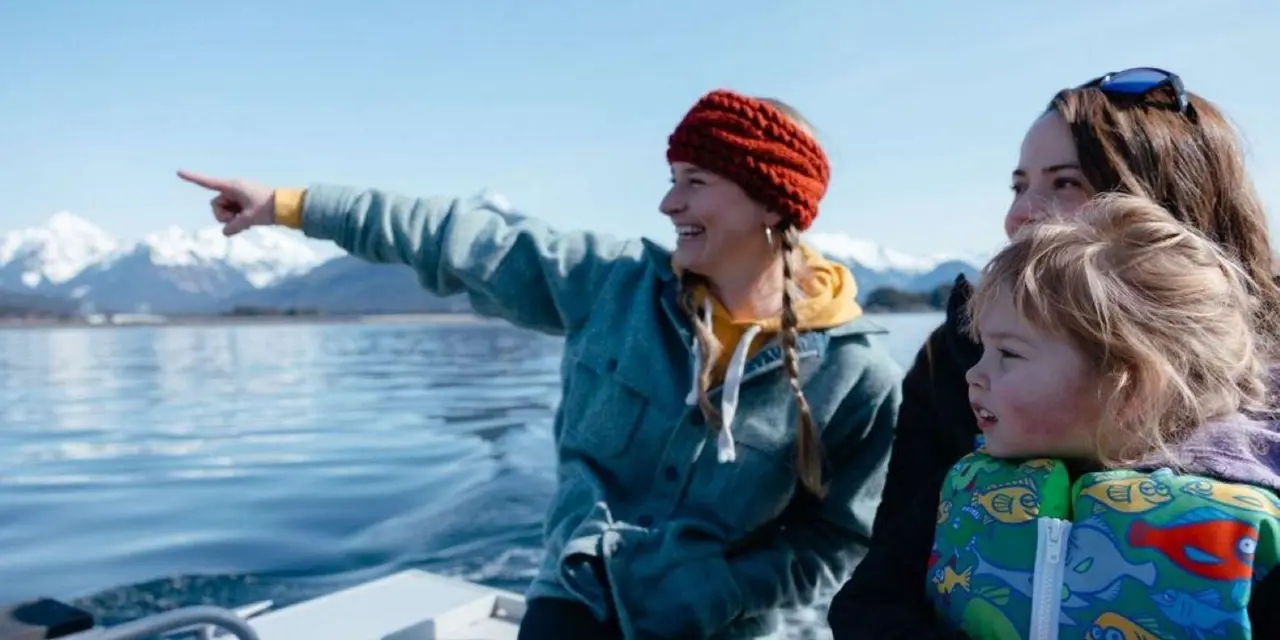 A smiling guide in a red knit headband pointing out across calm water from a small boat, with a child in a life jacket beside her and snow-capped peaks behind