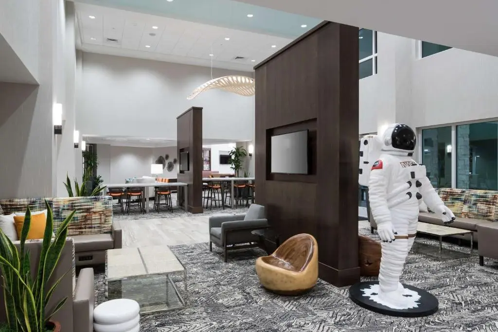The Hampton Inn & Suites lobby with grey sofas and high-top tables, and a life-size white astronaut statue standing beside a wood-panelled column