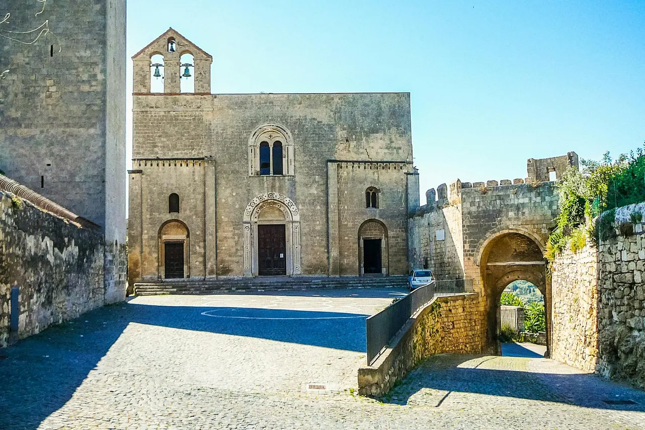 A medieval stone church with a bell gable and an arched town gate beside it under a clear blue sky
