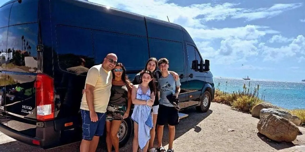 A family of five posing in front of a dark tour van beside the sea, with a sailing boat on the horizon