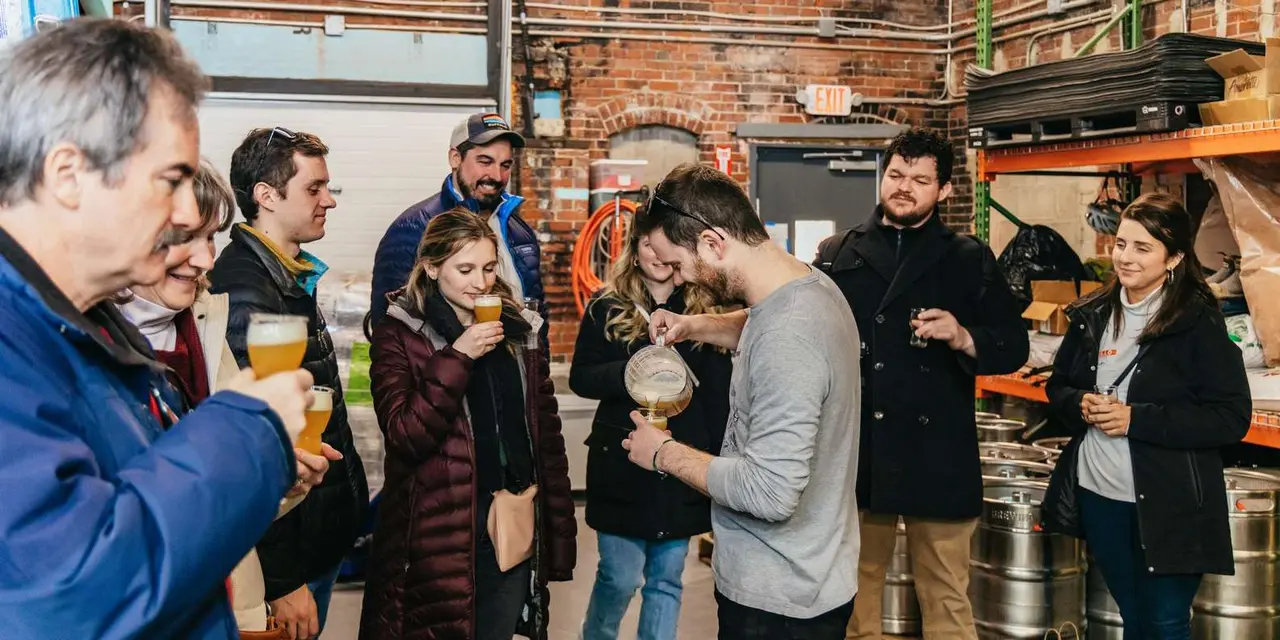 Guests sampling beer from glasses and a pitcher in a brick-walled brewery