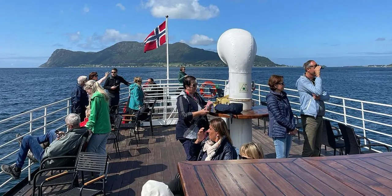 Passengers on a sunny boat deck with a Norwegian flag and a rocky island behind