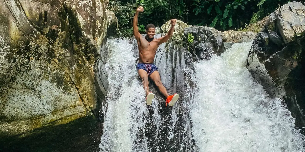A guide-led swimmer standing mid-jump on a rock beside a rushing rainforest waterfall