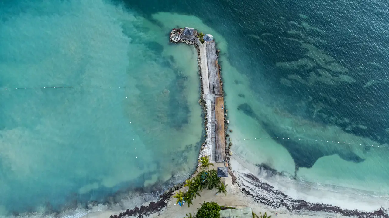 Aerial view of a stone jetty with a thatched gazebo at its tip, reaching into turquoise water off a palm-lined beach in Montego Bay, Jamaica
