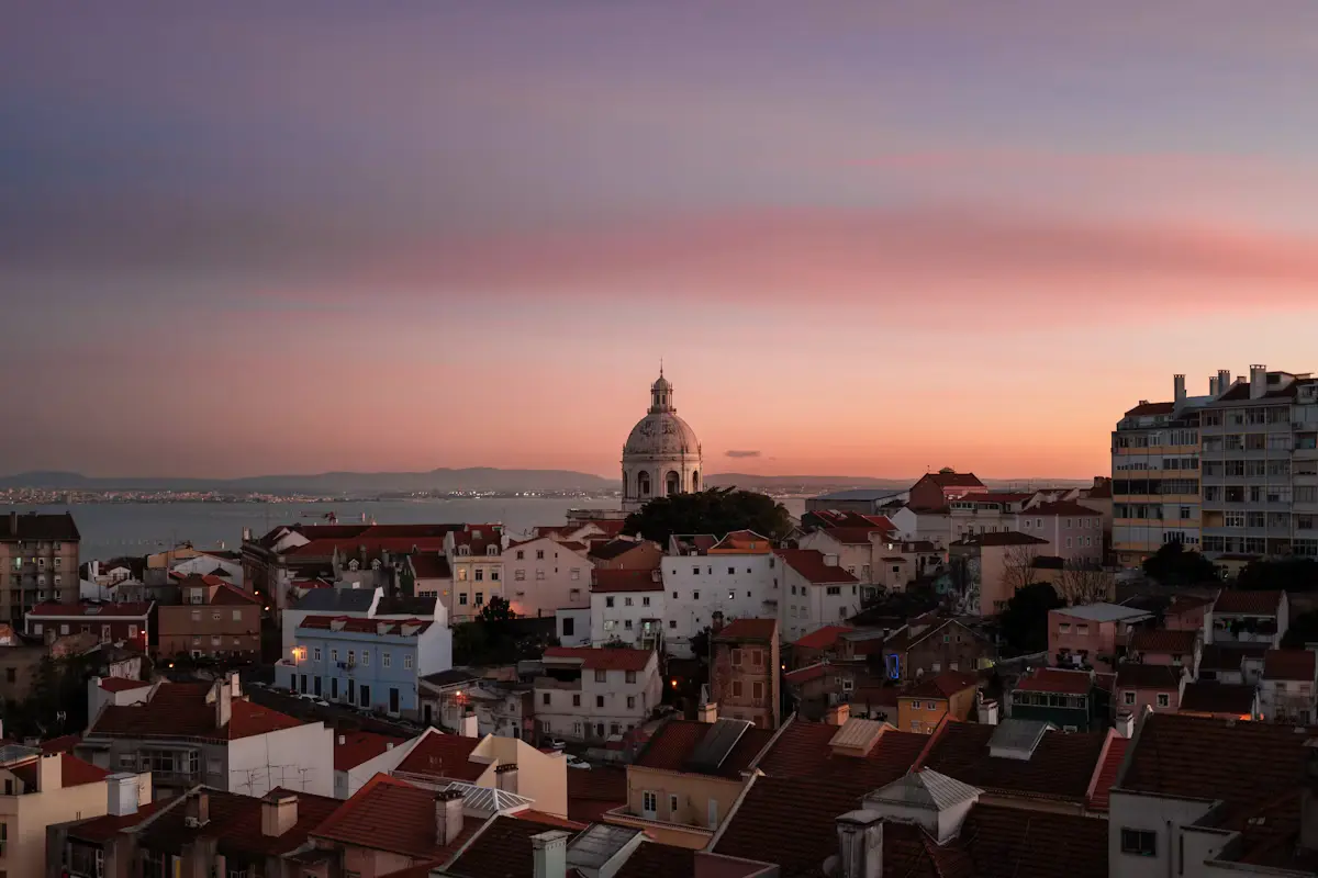 Dusk over Alfama's terracotta rooftops, with the dome of the National Pantheon and the Tagus estuary under a pink and purple sky