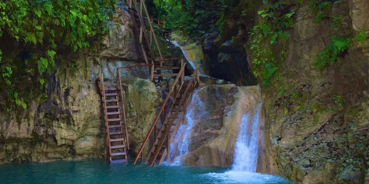 Wooden ladders climbing a rock wall beside twin waterfalls pouring into a turquoise pool