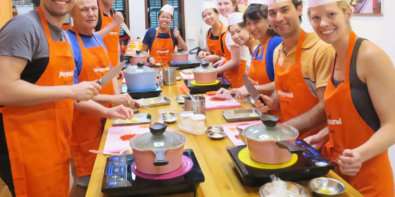 A class of cooks in orange aprons stirring pots on tabletop burners at a cooking studio