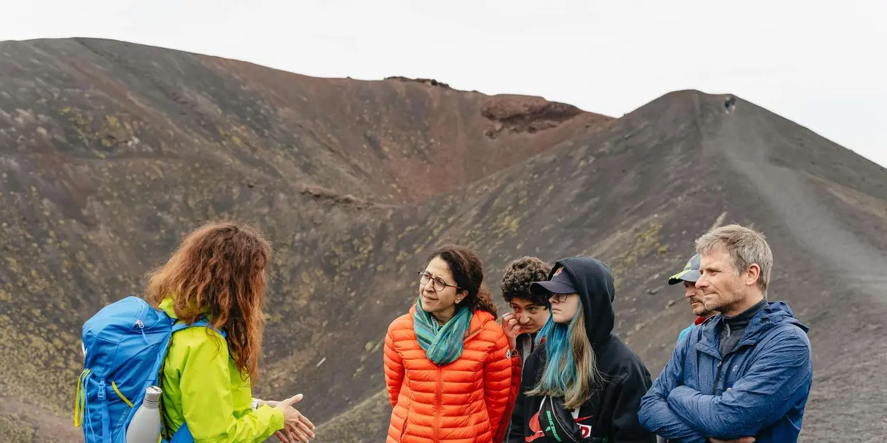 A guide talking to four hikers in jackets and beanies in front of a brown volcanic crater on Mount Etna