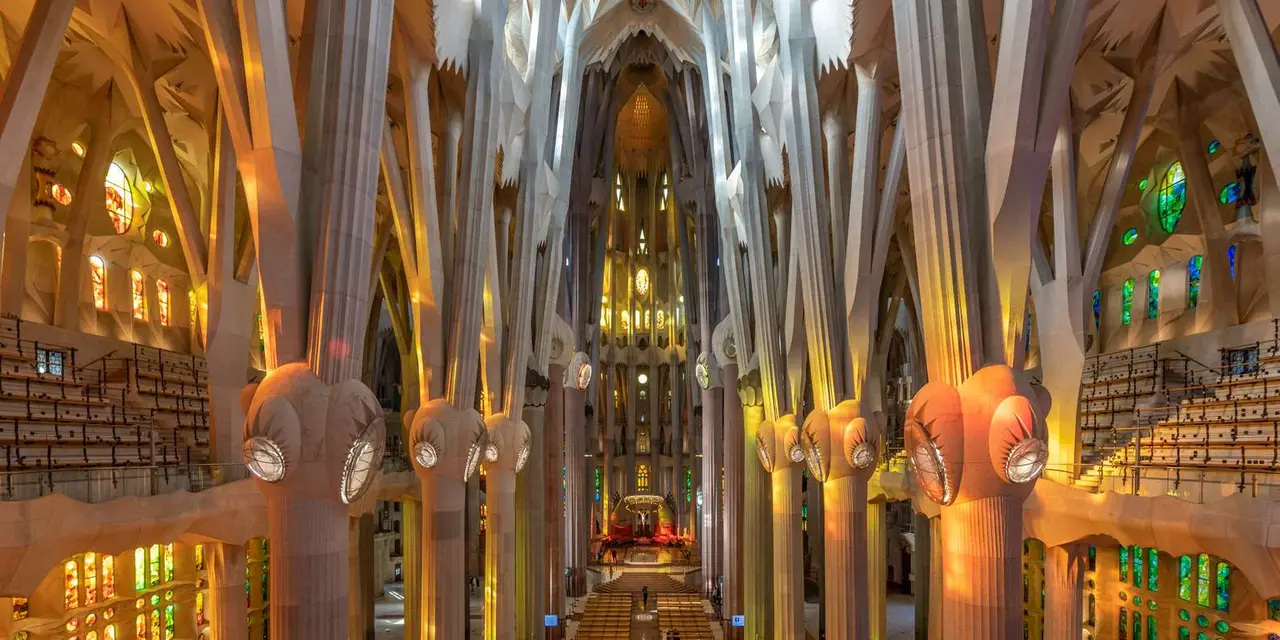 Looking down the nave of the Sagrada Família, tall tree-like columns branching toward a vaulted ceiling with stained glass glowing orange and green