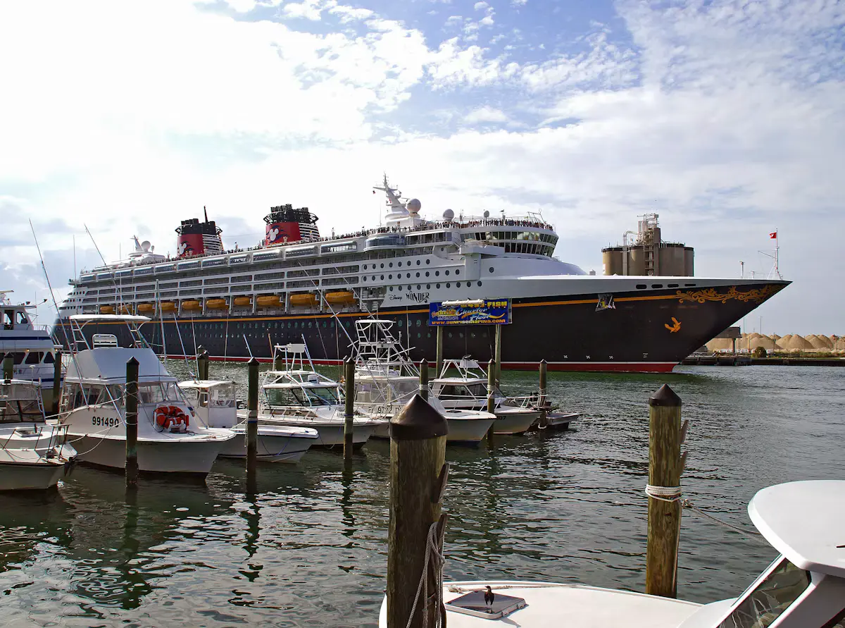 The Disney Wonder, black hull with a gold scroll bow and red funnels, sliding out of Port Canaveral past small fishing boats moored at wooden pilings, under a partly cloudy sky