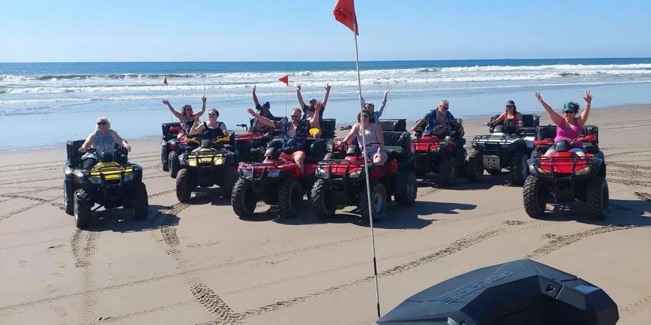 A line of riders on red quad bikes on a wide beach, several with arms raised, under a red flag